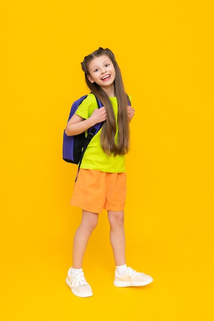 Une petite fille avec un sac à dos d'école Un enfant en short d'été va à des cours éducatifs Des cours supplémentaires Une écolière pleine longueur sur fond jaune isolé