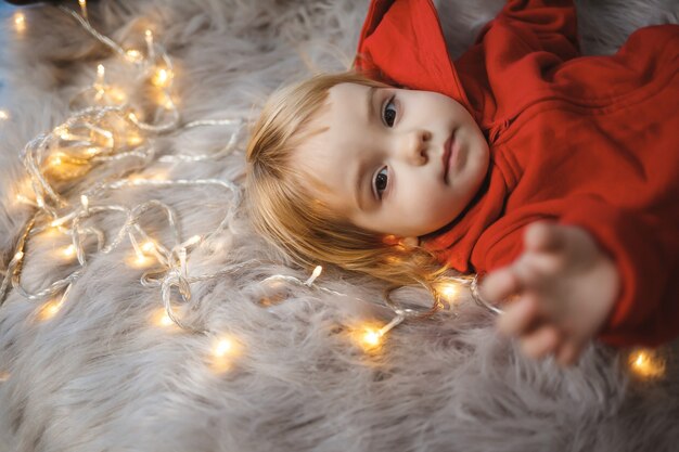 Petite fille en salopette de Noël d'humeur festive allongée sur le lit autour d'une guirlande lumineuse