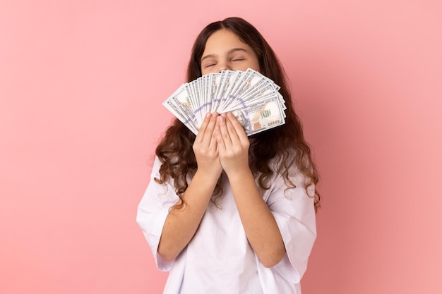 Petite fille satisfaite tenant un grand fan d'argent qui sent les billets en dollars garde les yeux fermés