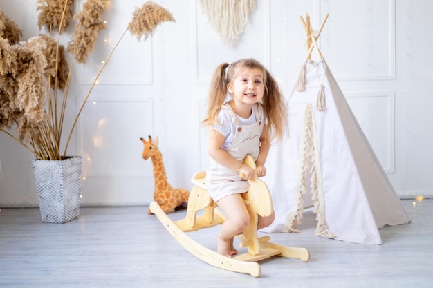 Une petite fille se balance sur un cheval à bascule en bois à la maison dans une pépinière lumineuse et rit