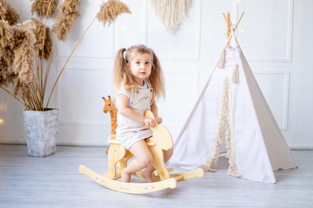 Une petite fille se balance sur un cheval à bascule en bois à la maison dans une pépinière lumineuse et rit