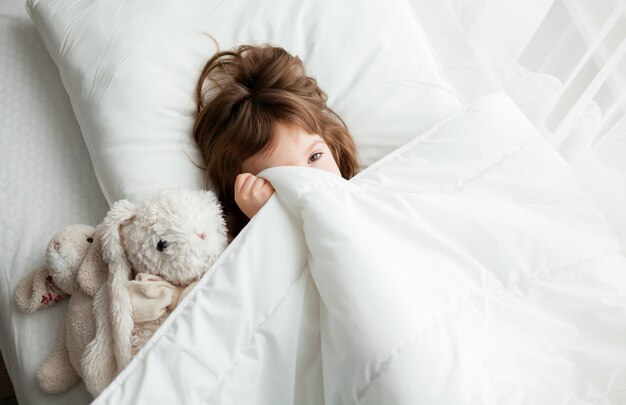 Petite fille se cachant sous une couverture blanche, elle vient de se réveiller