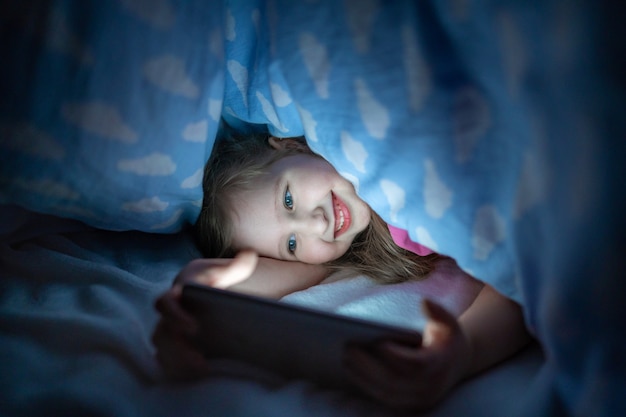 Petite fille se cache sous une couverture avec smartphone la nuit quand tout le monde dort.