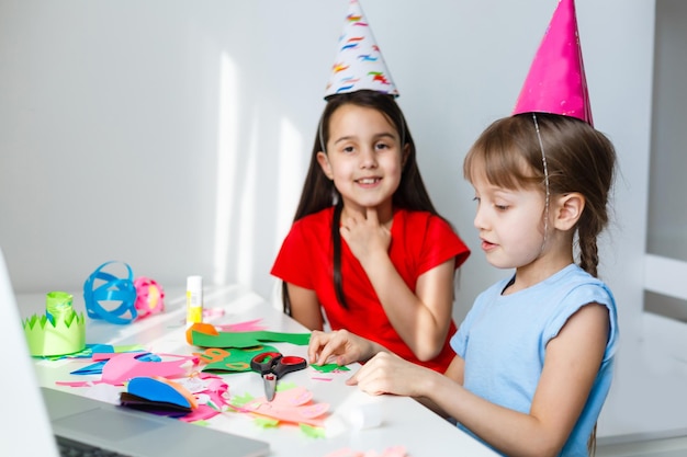 Une petite fille, se courbe et rit devant un ordinateur portable en casquette. Célèbre l'anniversaire via Internet en temps de quarantaine, l'auto-isolement et les valeurs familiales, l'anniversaire en ligne. Félicitations à l'animatrice