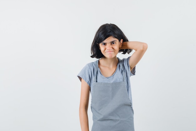 Petite fille se gratter la tête en t-shirt, tablier et à la honte, vue de face.