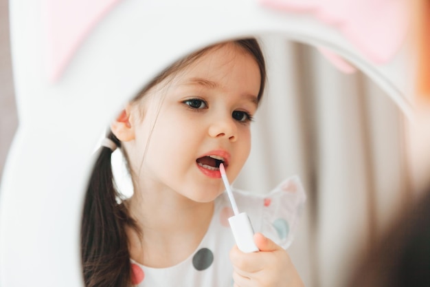 Une petite fille se regarde dans le miroir et peint ses lèvres avec des paillettes roses pour bébé