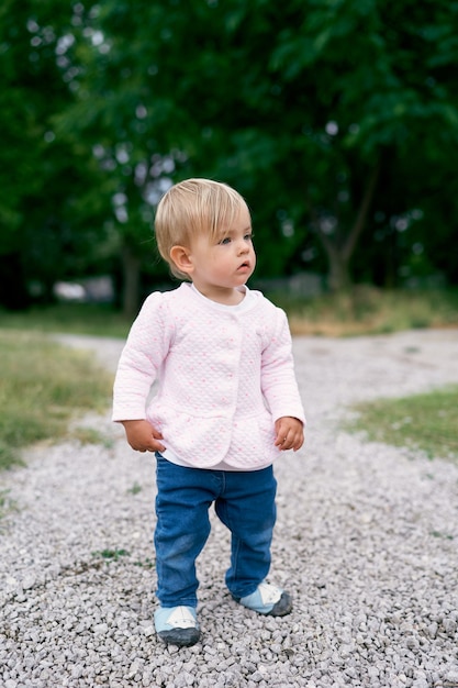 La petite fille se tient sur un chemin de gravier dans un parc verdoyant