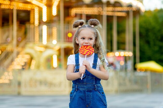 Une petite fille se tient dans un parc d'attractions avec une grande sucette