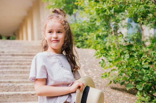 Une petite fille se tient dans le parc et tient un chapeau dans ses mains. L'enfant est heureux et rit.