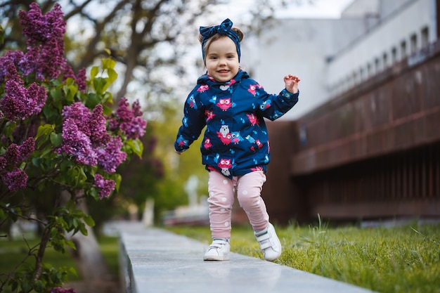 Une petite fille se tient près d'un buisson luxuriant de lilas, elle sourit et renifle des fleurs violettes