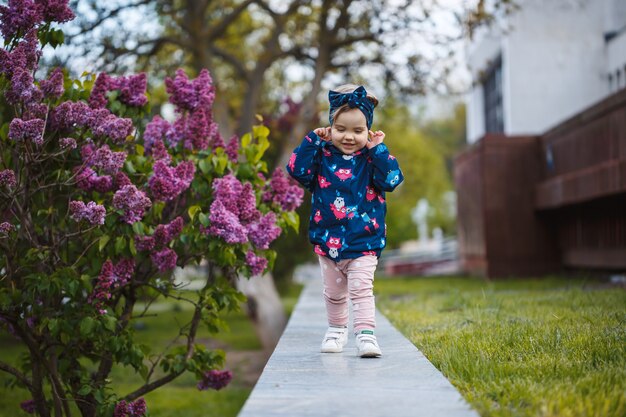 Une petite fille se tient près d'un buisson luxuriant de lilas, elle sourit et renifle des fleurs violettes