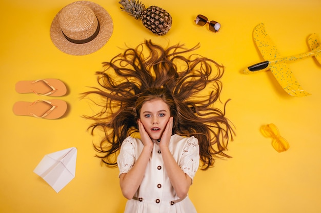 Photo une petite fille se trouve sur un fond jaune, et autour d'elle, des pantoufles, de l'ananas, un chapeau et un avion