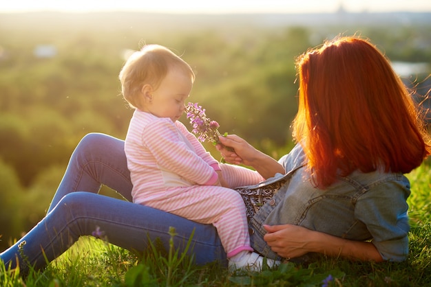 Petite fille sentant les fleurs assis sur maman, couché sur l&#39;herbe pendant le coucher du soleil.