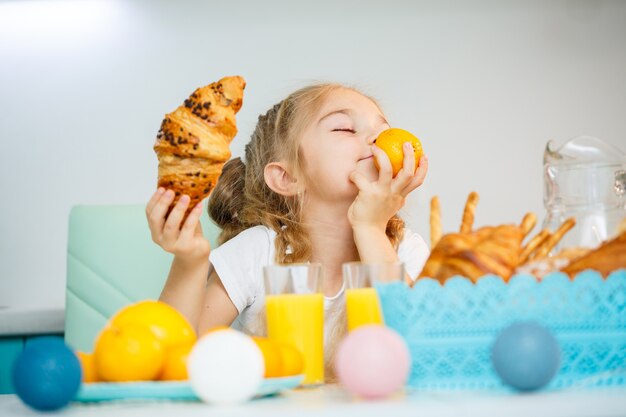 Une petite fille de sept ans, vêtue d'un tee-shirt blanc, est assise à la table de la cuisine. Contient des mandarines à la mandarine et un croissant fraîchement cuit au chocolat