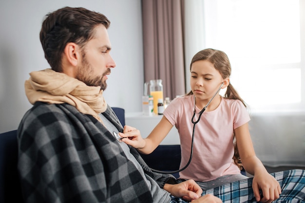 Une petite fille sérieuse écoute la respiration de papa à travers un stéthoscope. Un jeune homme malade s'assoit et ne bouge pas. Il regarde sa fille. Guy recouvert d'une couverture et d'un foulard dans la gorge.