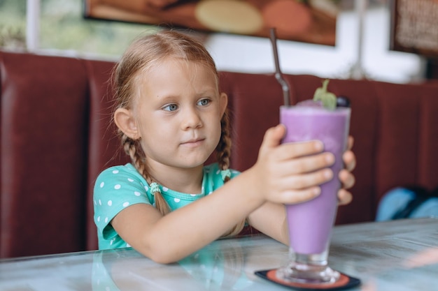 Une petite fille sérieuse avec des nattes blondes regarde un grand verre de milkshake et savoure un dessert sucré dans un café confortable local. Portrait.