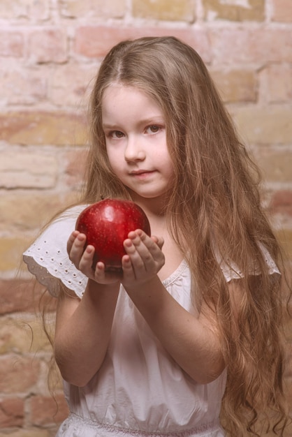 Petite fille sérieuse en robe blanche avec une grosse pomme rouge sur fond de mur de briques