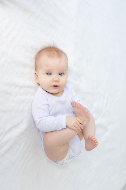 Petite fille de six mois joue avec ses pieds allongée sur le dos sur un lit en coton blanc sur un lit dans la chambre de la maison