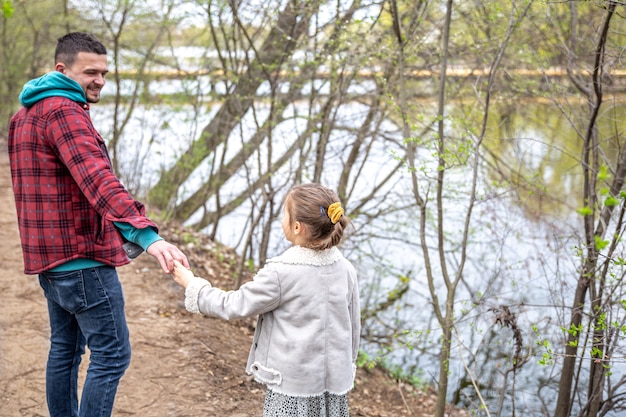 Une petite fille avec son père au début du printemps, par temps froid, marche près de la rivière, se tenant la main.