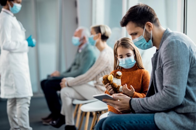 Petite fille et son père avec des masques faciaux à l'aide d'un pavé tactile tout en étant assis dans la salle d'attente d'une clinique médicale