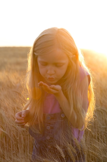 Petite fille soufflant sur ses paumes remplies de grains de blé debout au champ de blé au coucher du soleil