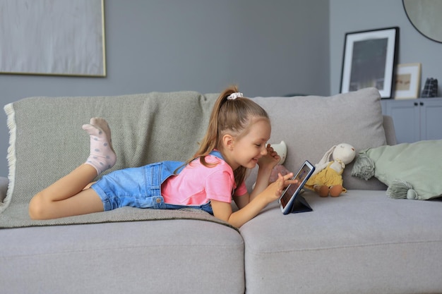 Petite fille souriante allongée sur un canapé jouant à des jeux en ligne, surfant sur le Web, utilisant des applications amusantes sur tablette.