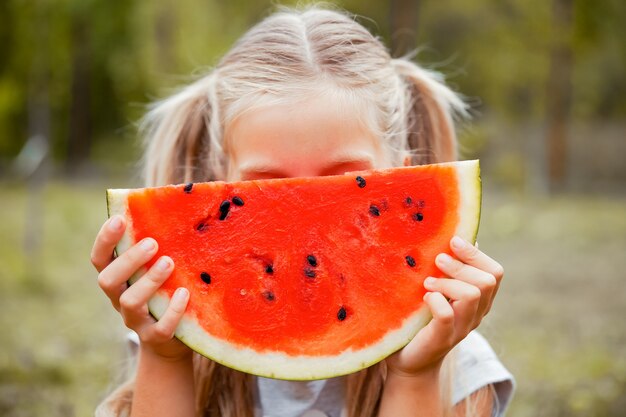 Petite fille souriante aux yeux bleus mangeant un morceau de pastèque à l'extérieur.