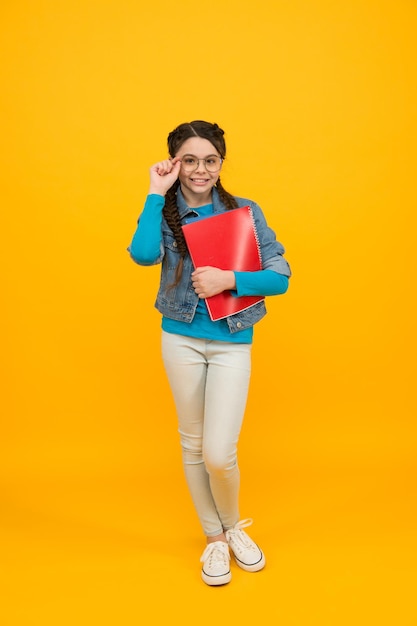 Une petite fille souriante avec des cheveux tressés élégants portant des lunettes tient un cahier de travail sur fond jaune de retour à l'école