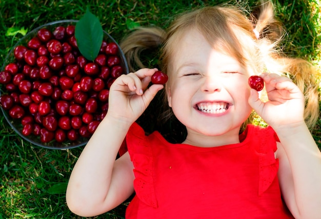 petite fille souriante dans une chemise rouge allongée sur l'herbe près d'un bol de cerises tenant des cerises dans les mains