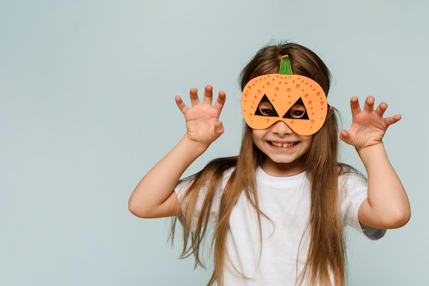 Une petite fille souriante dans un masque de citrouille célèbre Halloween sur fond bleu