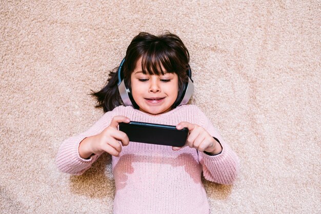 Petite fille souriante dans un pull rose allongé sur un tapis écoutant de la musique et regardant des films sur son téléphone portable portant des écouteurs sans fil Concept de technologie petits enfants et mobiles