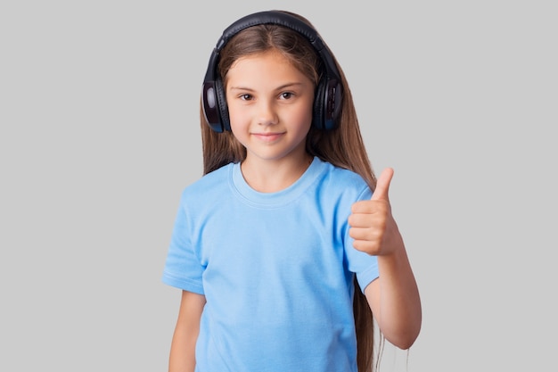 Petite fille souriante écoutant de la musique et montrant les pouces vers le haut sur un fond gris