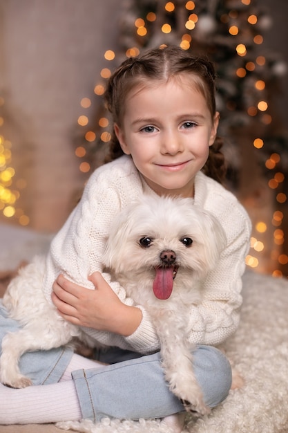 Une petite fille souriante embrasse un chien maltais sur le fond de Noël des lumières à la maison