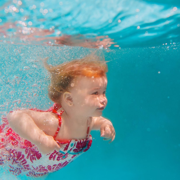 Petite fille souriante en jolie robe moderne plongeant sous l'eau dans la piscine bleue
