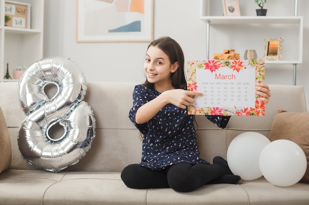 Petite fille souriante le jour de la femme heureuse tenant et points au calendrier assis sur un canapé dans le salon