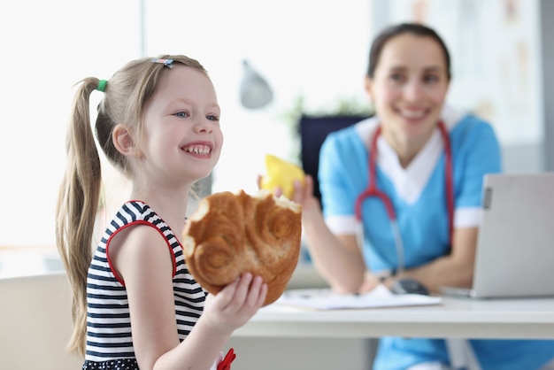 Une petite fille souriante mange un petit pain sur fond de docteur