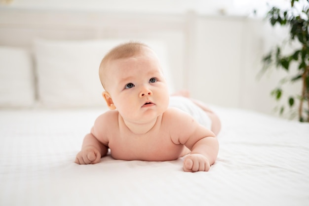 Une petite fille souriante mignonne en culotte blanche est allongée sur le lit dans la chambre de la maison sur une literie blanche sur le ventre en regardant la caméra en souriant bébé heureux