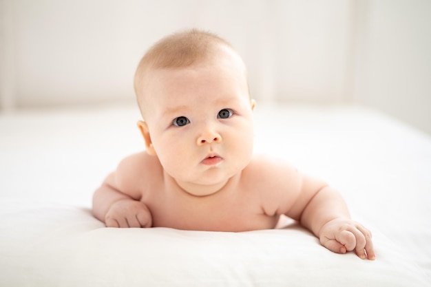 Une petite fille souriante mignonne en culotte blanche est allongée sur le lit dans la chambre de la maison sur une literie blanche sur le ventre en regardant la caméra en souriant bébé heureux