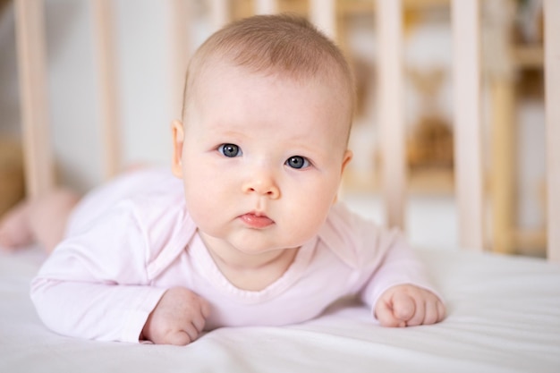 Une petite fille souriante mignonne dans un body est allongée dans un berceau à la maison sur une literie blanche sur le ventre en regardant la caméra en souriant un bébé heureux