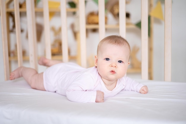 Une petite fille souriante mignonne dans un body est allongée dans un berceau à la maison sur une literie blanche sur le ventre en regardant la caméra en souriant un bébé heureux