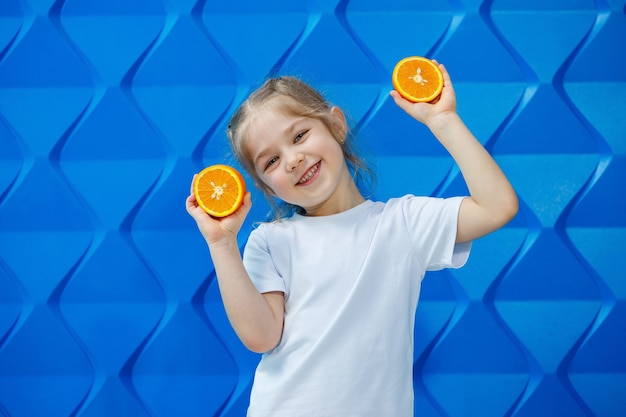 Petite fille souriante avec des nattes dans un T-shirt blanc sur fond bleu avec une orange coupée dans ses mains. Émotions des enfants, amusement