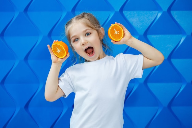 Petite fille souriante avec des nattes dans un T-shirt blanc sur fond bleu avec une orange coupée dans ses mains. Émotions des enfants, amusement