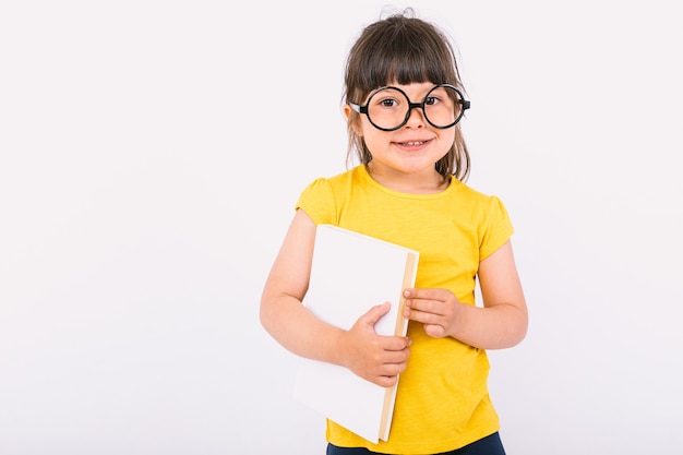 Petite fille souriante portant un t-shirt jaune et des lunettes noires rondes tenant un livre dans ses mains sur fond blanc