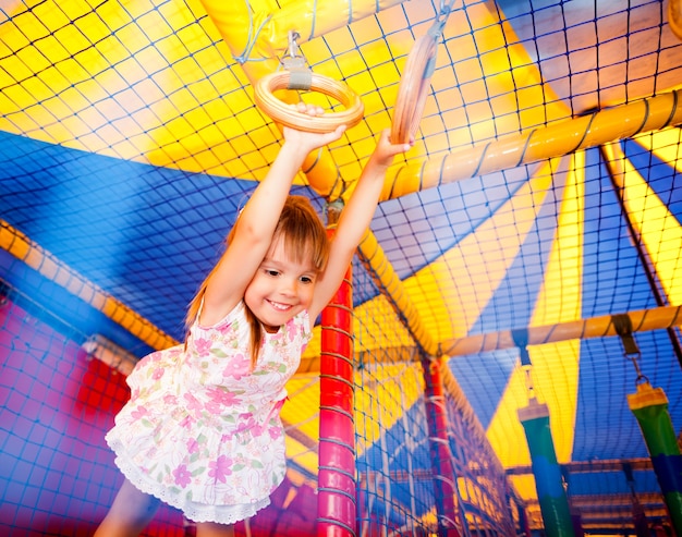 Petite fille souriante en robe accrochée à des anneaux et se sentir excité dans la salle de jeux.