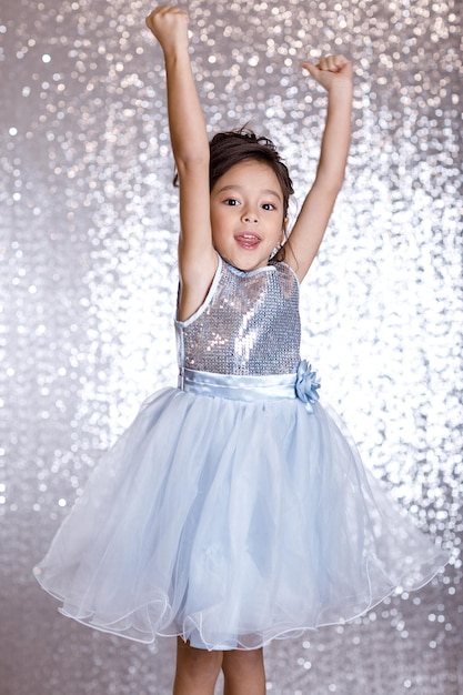 Petite fille souriante en robe argentée et bleue sautant sur fond avec bokeh argenté. fête d'anniversaire