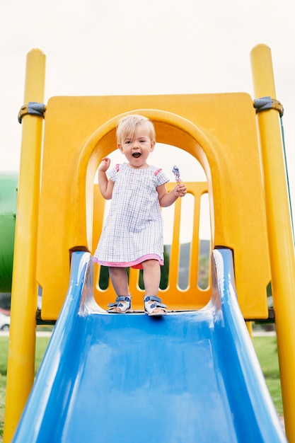 Une petite fille souriante se dresse sur le dessus du toboggan sur le terrain de jeu