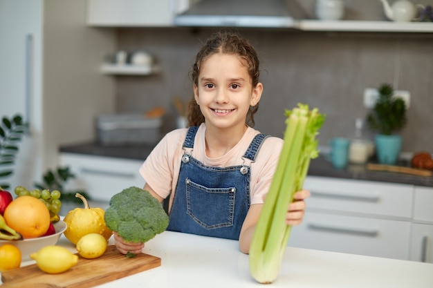 Une petite fille souriante se tient près de la table dans la cuisine, tenant du brocoli vert et du céleri dans les mains, regardant la caméra. Vue horizontale.