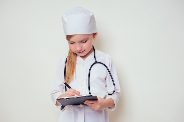 Petite fille souriante en uniforme de médecin avec stéthoscope d'outils médicaux écrivant quelque chose dans le presse-papiers sur fond blanc dans l'espace de copie de studio. future carrière d'orientation professionnelle