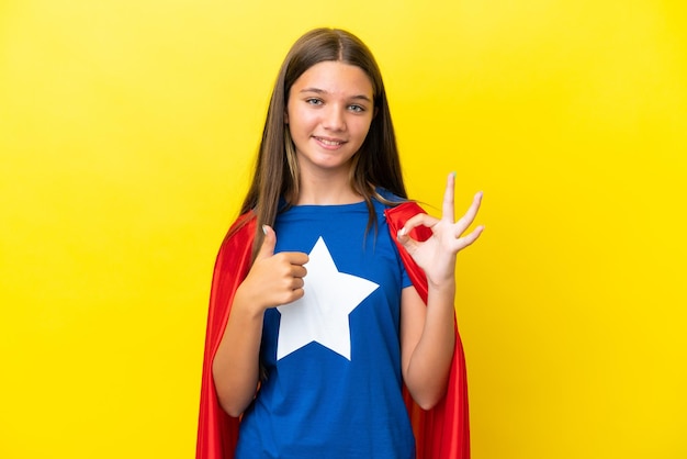 Petite fille de super-héros caucasien isolée sur fond jaune montrant le signe ok et le geste du pouce vers le haut