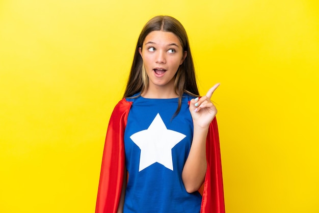 Petite fille de super-héros caucasien isolée sur fond jaune pensant à une idée pointant le doigt vers le haut